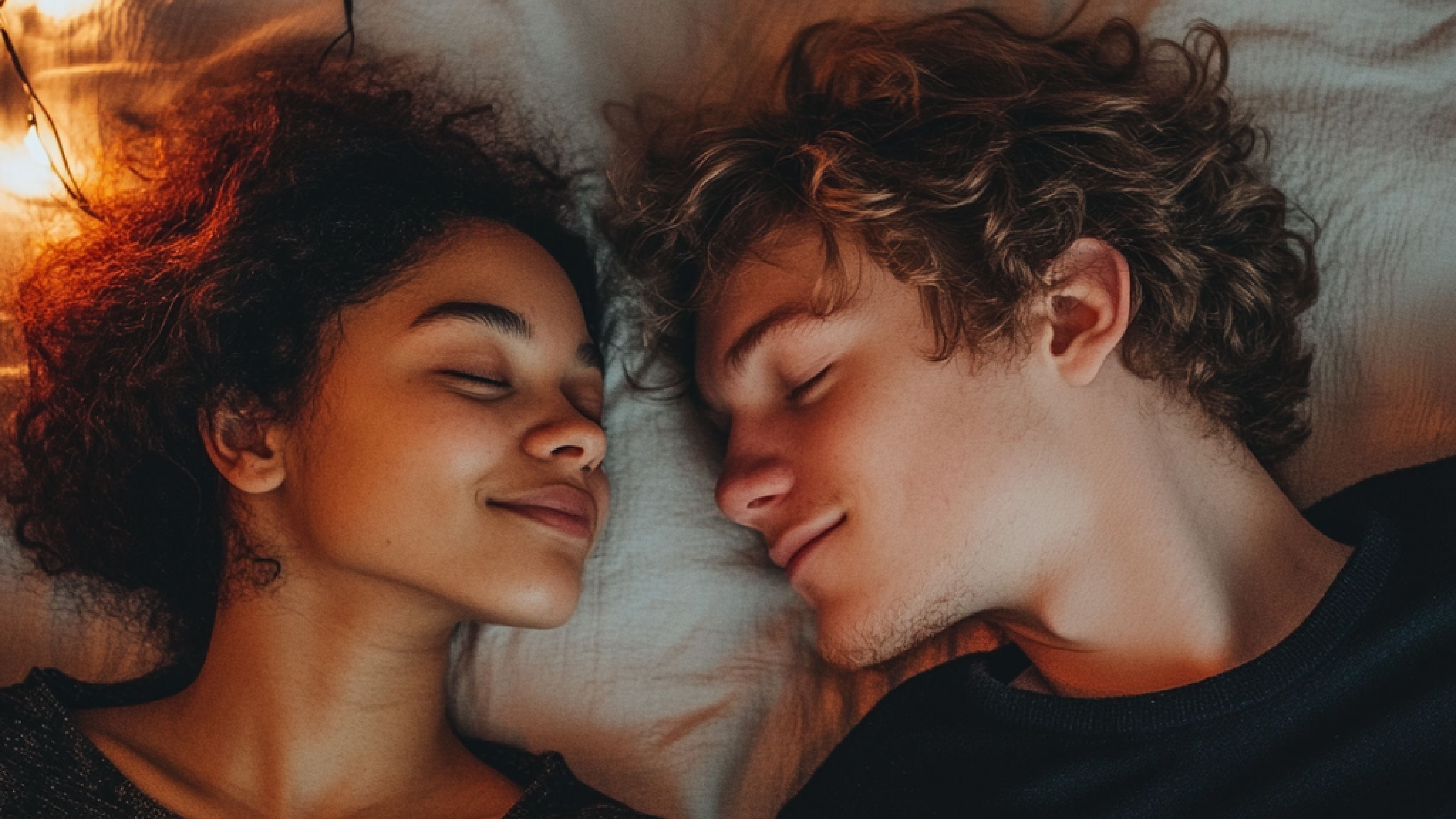 A close-up of a couple lying side by side, smiling with their eyes closed, on a soft bed.